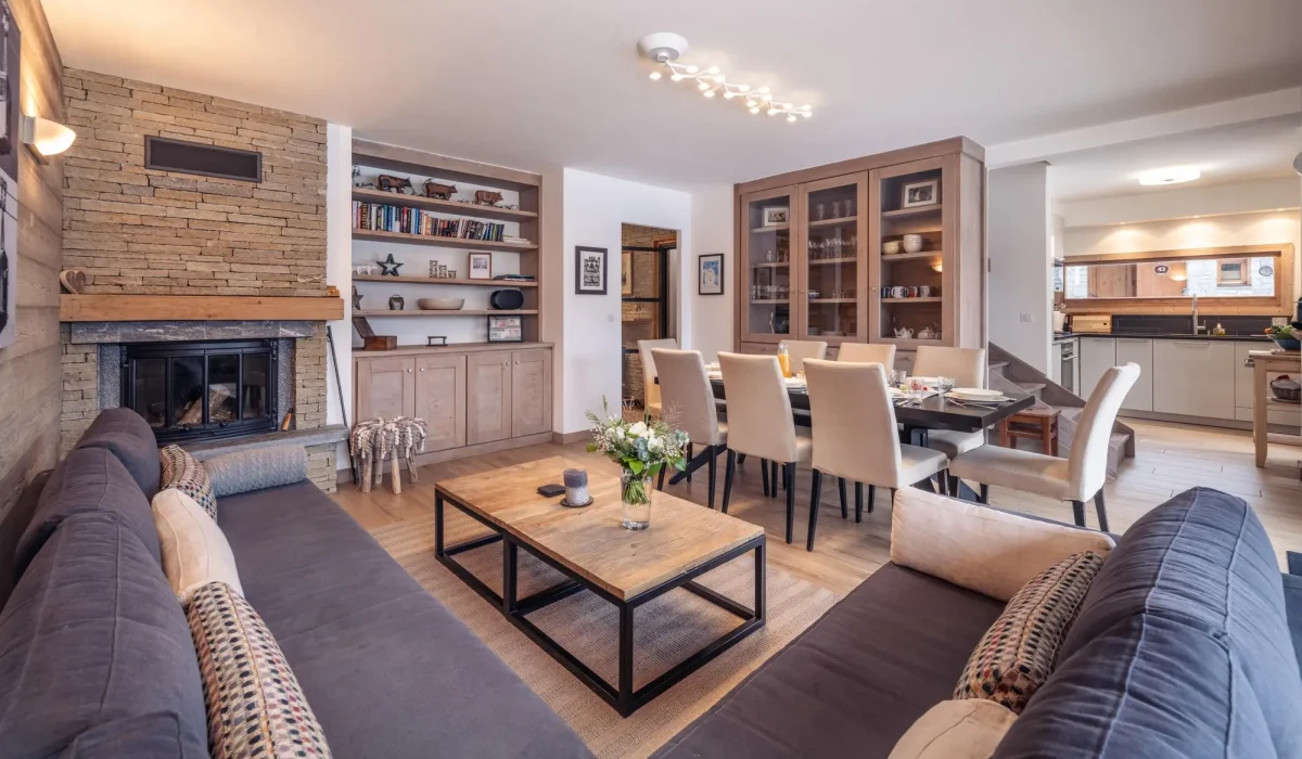Lounge with a stone fireplace, grey sofas and the dining room beyond at Chalet Alisier in Méribel Village