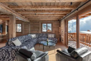 Lounge under reclaimed-wood beams with a corner sofa, leather armchairs and a glazed door onto the balcony at Chalet Arnica in Les Saisies