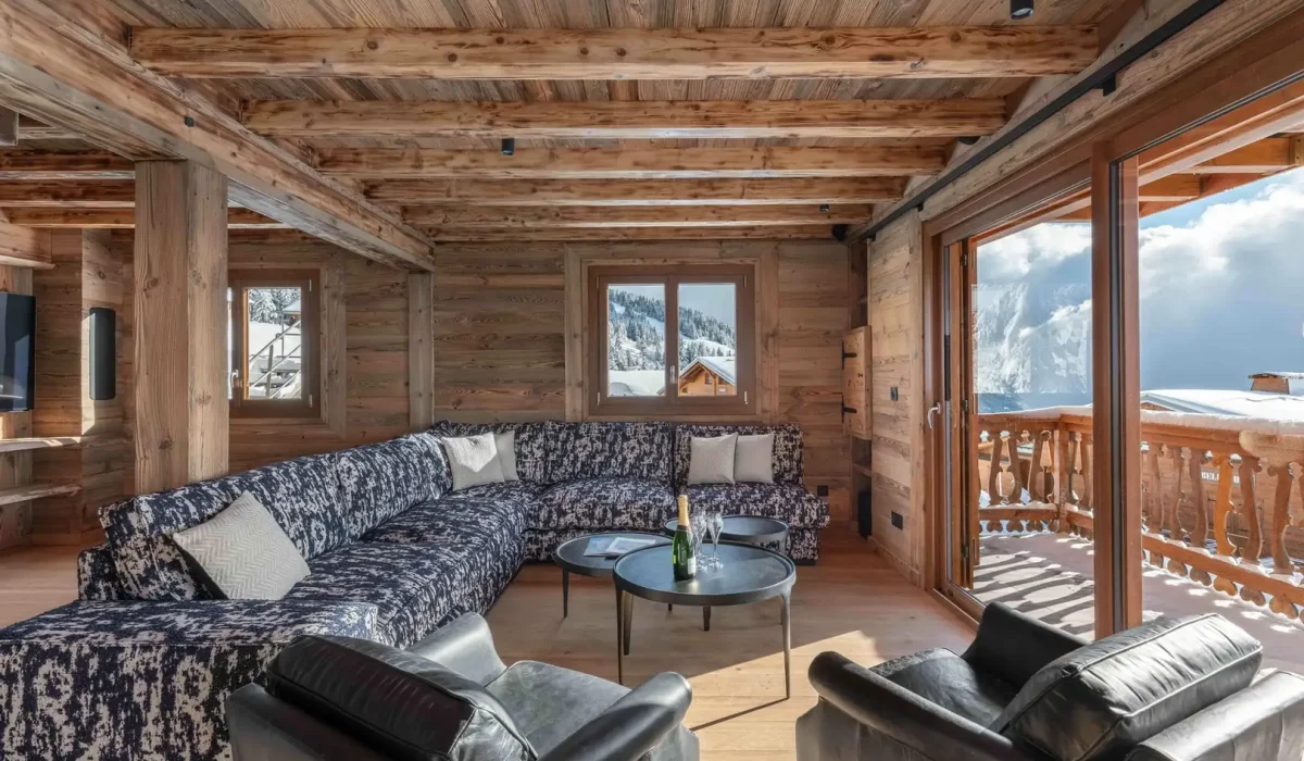 Lounge under reclaimed-wood beams with a corner sofa, leather armchairs and a glazed door onto the balcony at Chalet Arnica in Les Saisies
