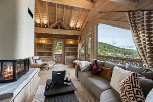 Lounge with a grey sofa, a central fireplace and a large picture window over the valley at Chalet Églantine in Courchevel La Tania
