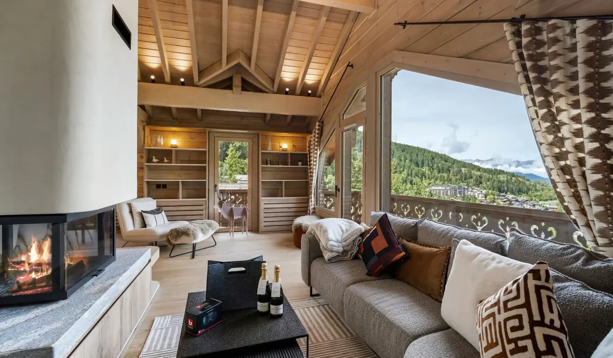 Lounge with a grey sofa, a central fireplace and a large picture window over the valley at Chalet Églantine in Courchevel La Tania