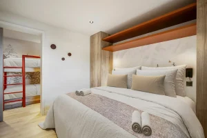 Double bedroom with a light wood headboard, orange shelving and bunk beds beyond at Chalet Églantine in Courchevel La Tania