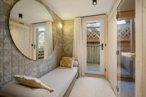 Relaxation room with a daybed, a large round mirror, the sauna door and access to the terrace at Chalet Églantine in Courchevel La Tania