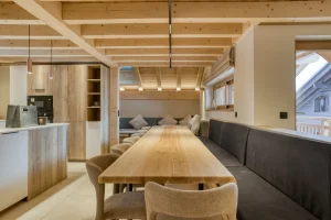 Living area with a long wooden table, a grey banquette and the open kitchen at Chalet Épicéa in Les Saisies