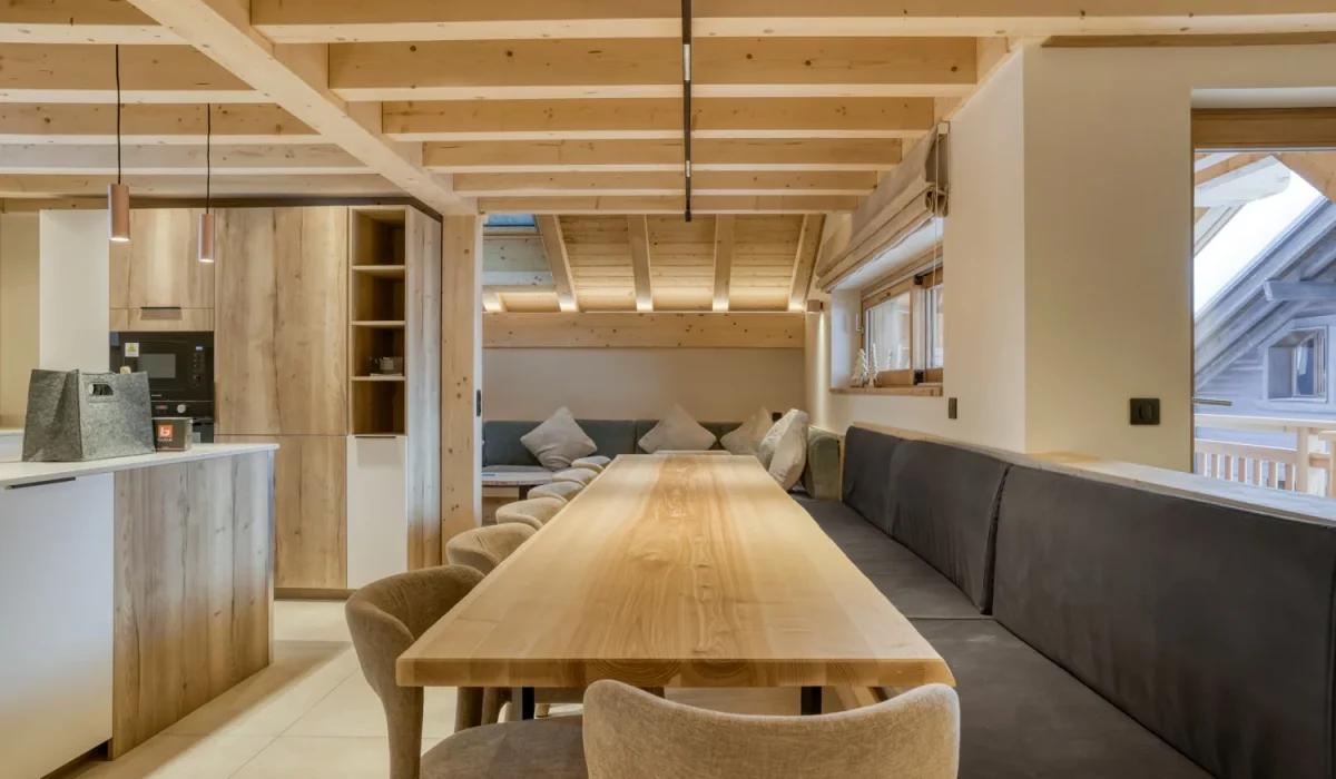 Living area with a long wooden table, a grey banquette and the open kitchen at Chalet Épicéa in Les Saisies
