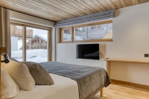 Bedroom with a bed, a wall-mounted TV and windows onto the neighbouring chalets at Chalet Épicéa in Les Saisies