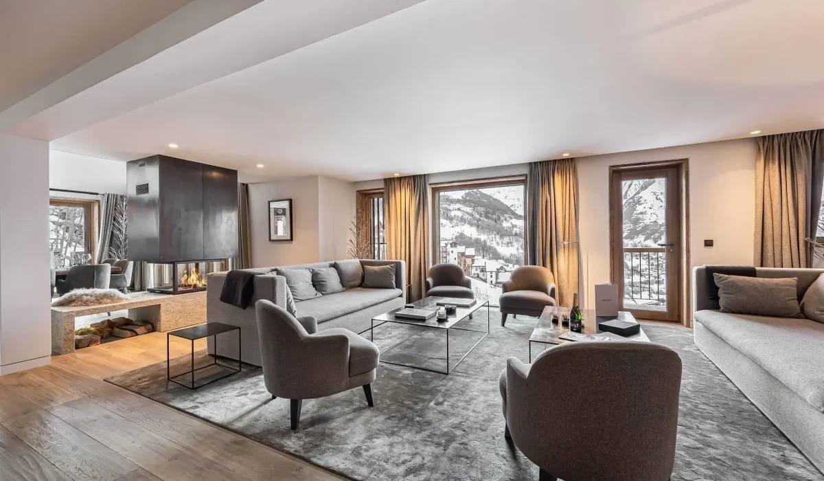 Living area with a lounge, grey armchairs and a central fireplace opening onto the large windows at Chalet Merlin in Saint-Martin-de-Belleville